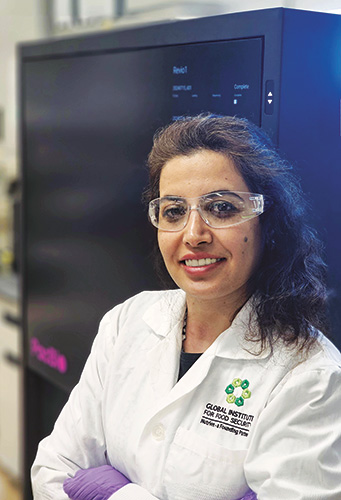 Hakimeh Emamgholi Begli, a quantitative geneticist, wears a lab coat, gloves, and safety goggles, and stands in front of a large rectangular piece of scientific equipment.