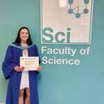 Ashlyn Kirk wears a graduation gown and stands holding her diploma beside the Faculty of Science sign.