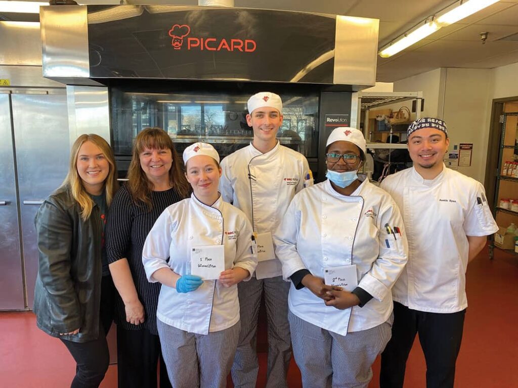 Two women stand with the four winners of RRC's scholarship. The winners are in their chef outfits and everyone is in an industrial kitchen.