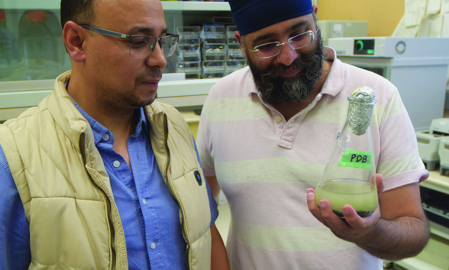 Harmeet Chawla and Mohamed Youssef together in a science lab. Harmeet holds a beaker that has the label PDB on it.