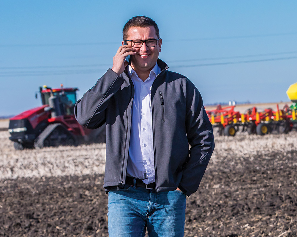 An agronomist is on his cellphone in a field, with farm equipment in the background.